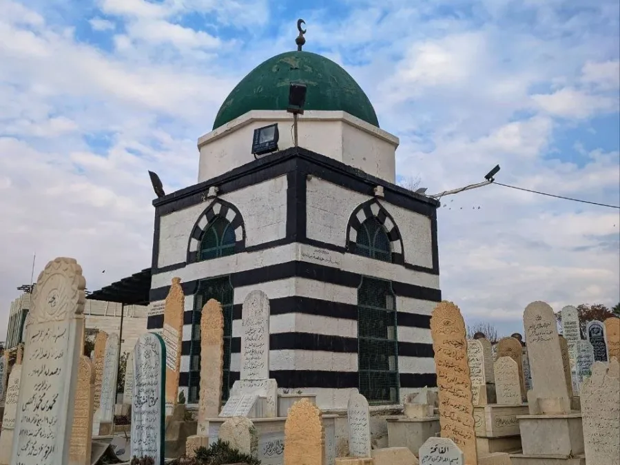 A green domed tomb in banded black and white stone standing among the carved headstones of the Bab al-Saghir cemetery in Damascus