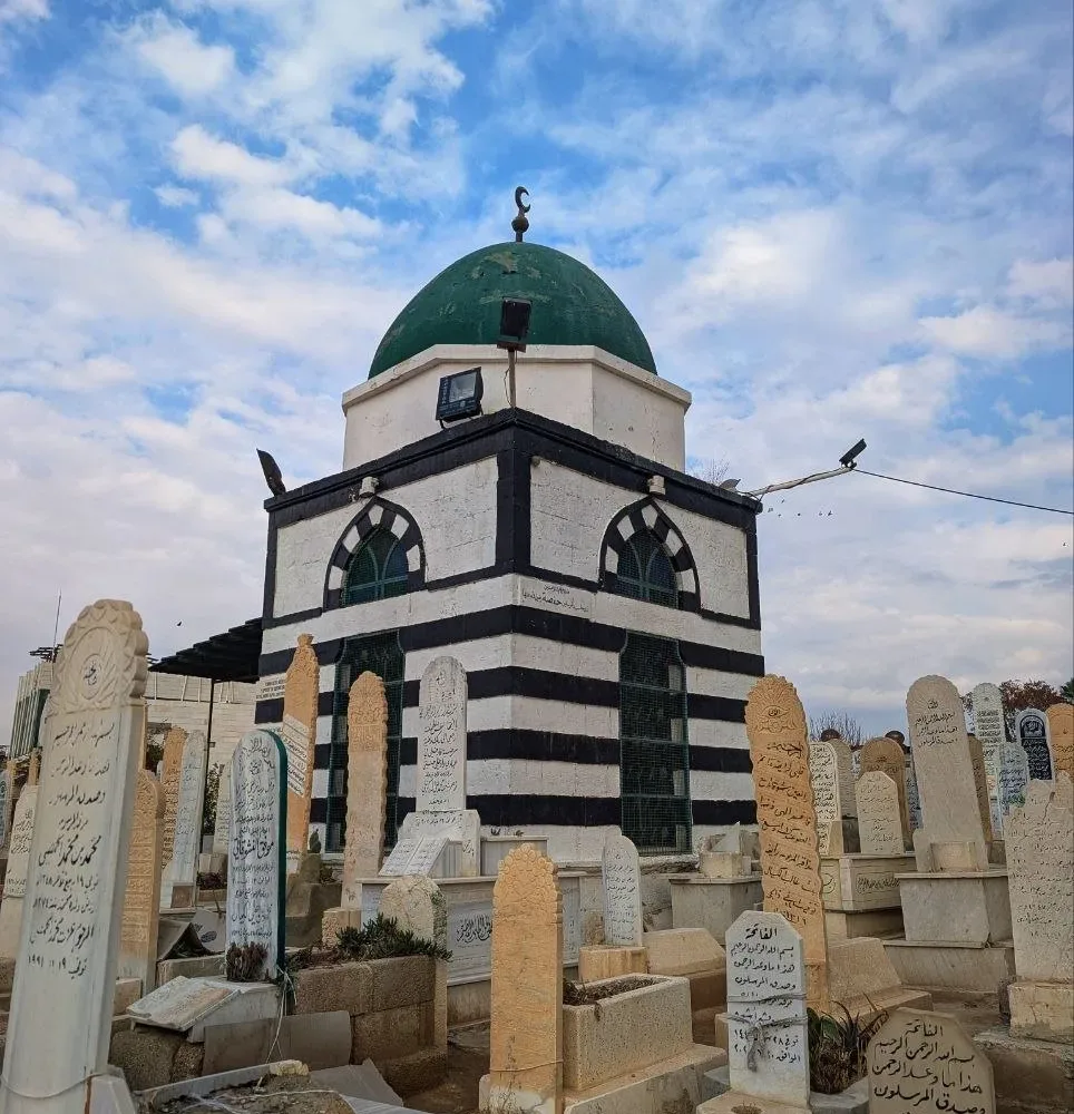 A green domed tomb in banded black and white stone standing among the carved headstones of the Bab al-Saghir cemetery in Damascus