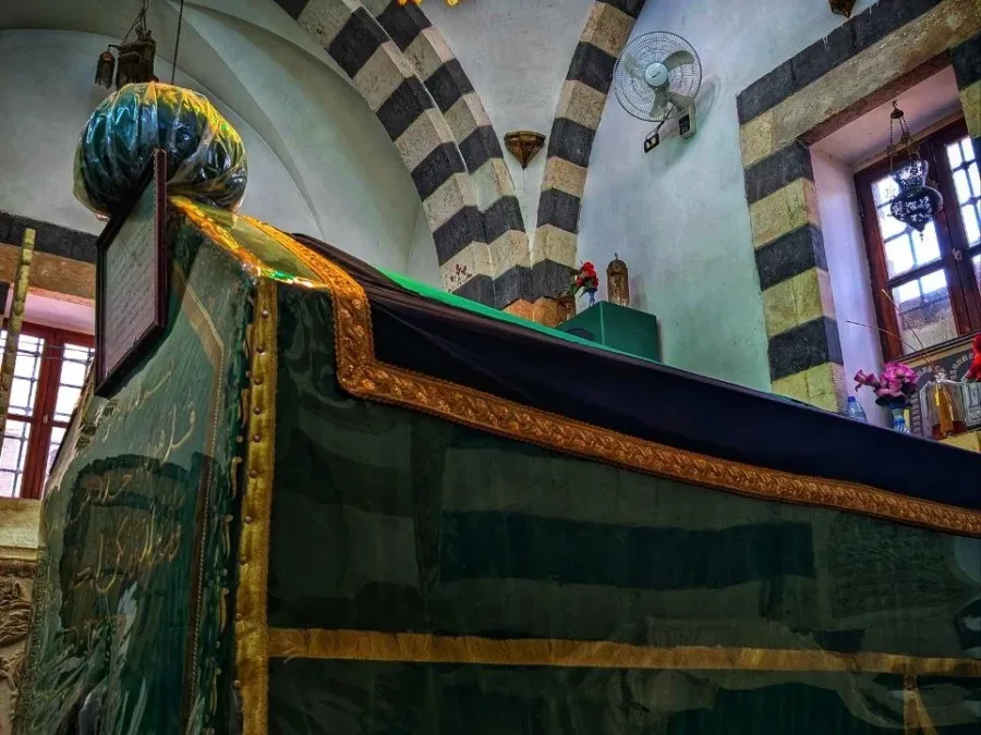 The green and gold draped tomb of Sayyidna Bilal beneath banded stone arches and a colored glass chandelier in Bab al-Saghir, Damascus