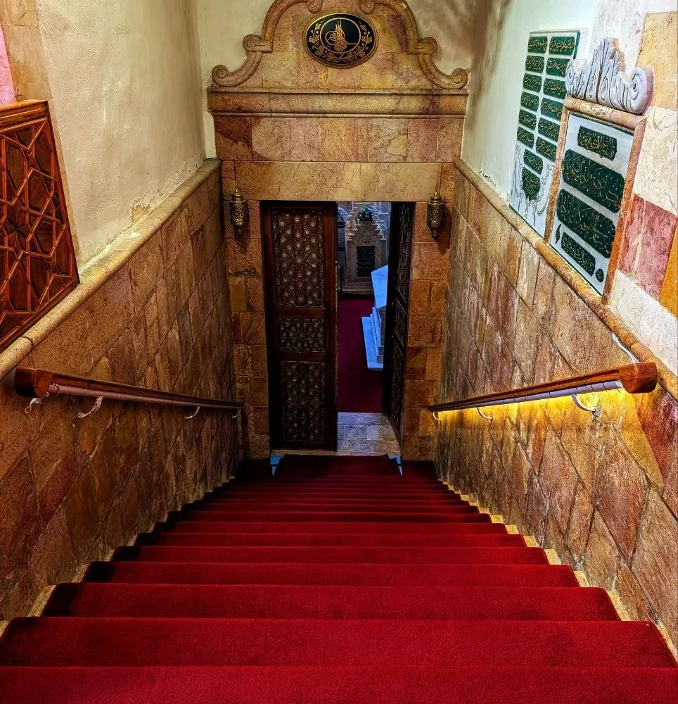 Red carpeted stairs leading down to the doorway of the tomb chamber of Ibn Arabi in Damascus, an Ottoman tughra carved on the marble above it