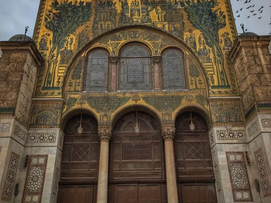 The gold and green mosaic gable of the Umayyad Mosque prayer hall in Damascus, birds wheeling over it against a gray sky