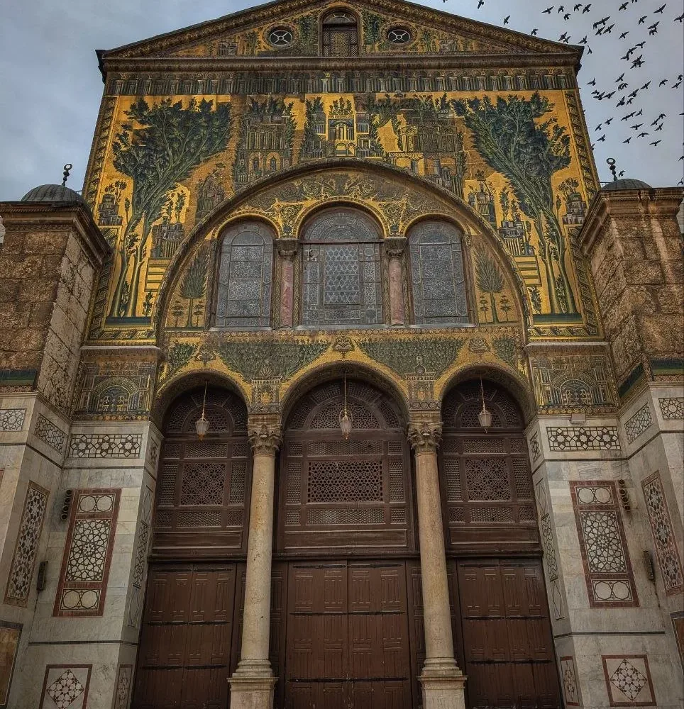 The gold and green mosaic gable of the Umayyad Mosque prayer hall in Damascus, birds wheeling over it against a gray sky